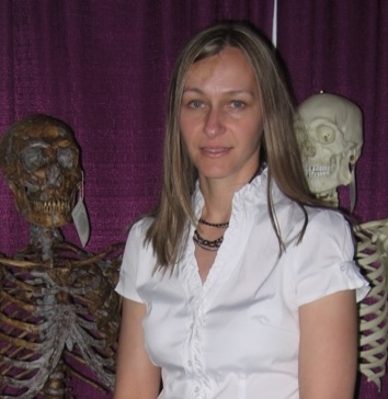 Woman with long light-brown hair in a white shirt, standing between two skeletons.